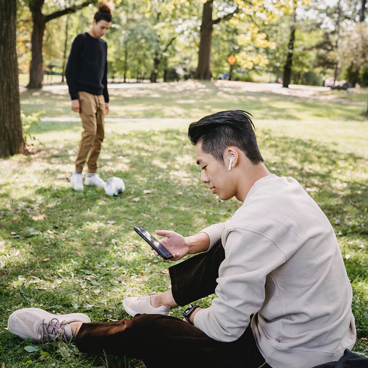 two men in a park with one kicking a soccer ball and the other sitting with headphones in looking at his phone